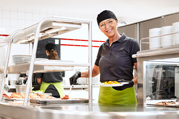 Employé de Dussmann, issu du secteur de la restauration et de l'hôtellerie, servant des repas dans une cafétéria avec le sourire