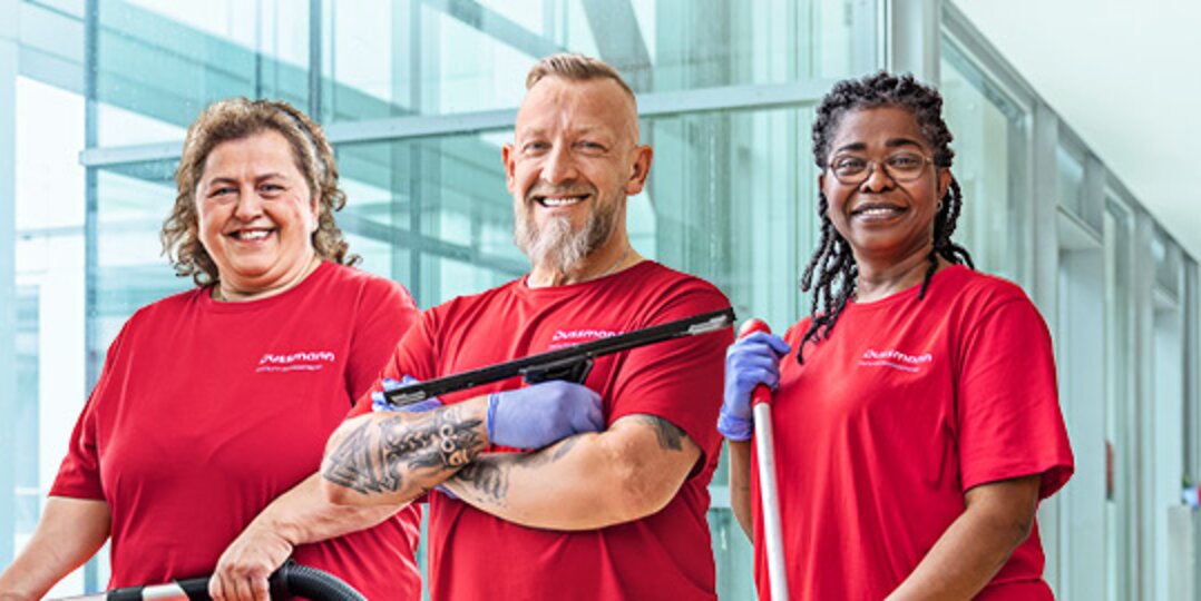 Photo de groupe de trois employés souriants de Dussmann, issus du secteur des services de nettoyage, devant un fond bleu