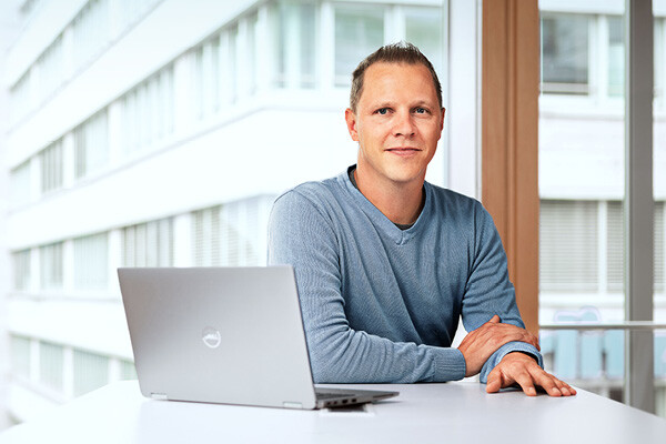 Employé de Dussmann, issu du secteur des services commerciaux, assis dans un bureau devant son ordinateur portable, affichant un sourire accueillant