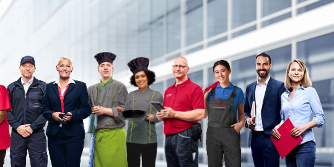 Photo de groupe d'employés souriants de Dussmann issus de différents domaines professionnels, devant un fond gris neutre