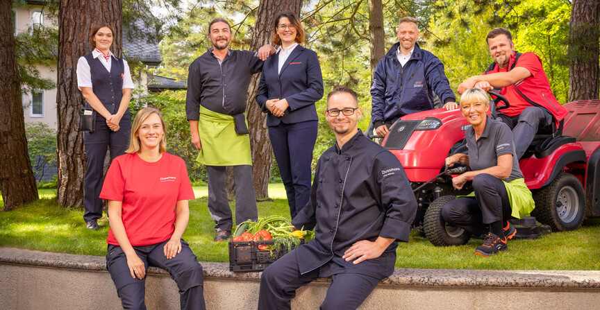Photo de groupe des employés souriants de Dussmann, issus de différents secteurs d'activité, debout dans une grande cour avec pelouse et arbres, vêtus de leurs tenues de travail